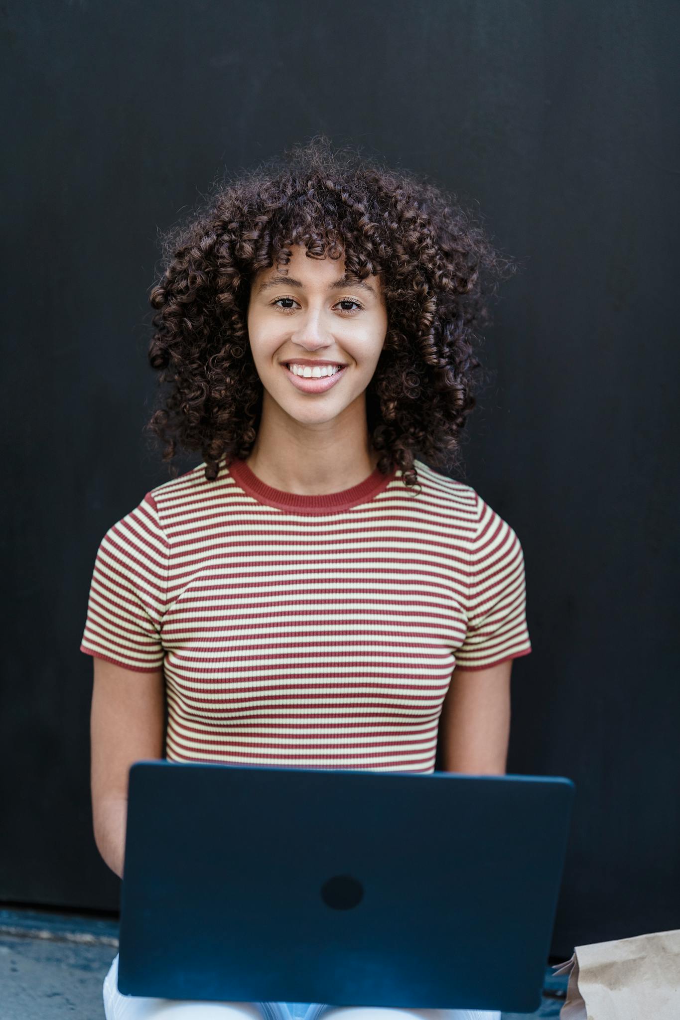 Cheerful young ethnic female student with dark curly hair in striped t shirt smiling and looking at camera while sitting on step on street and doing homework assignment on laptop
