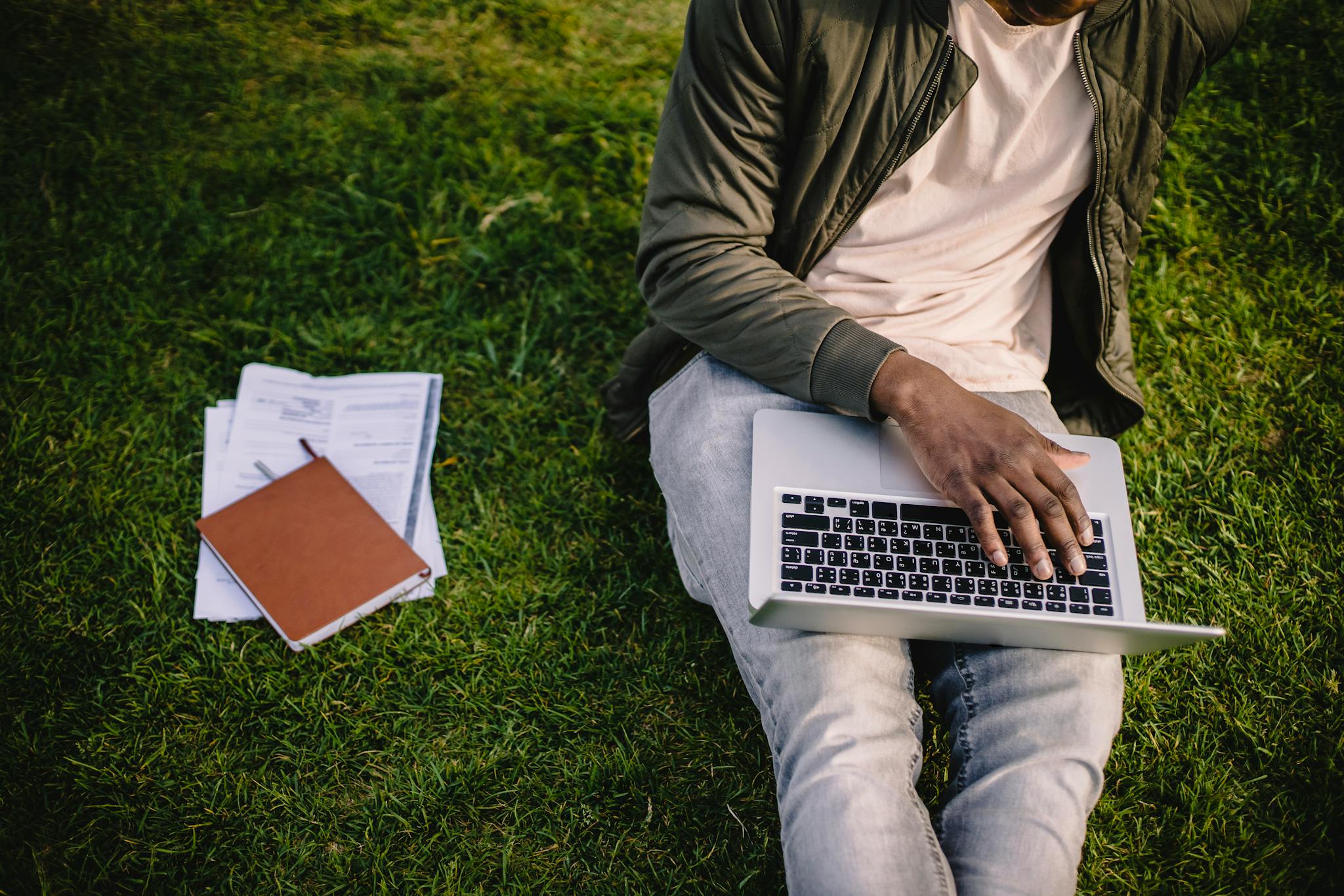 Crop unrecognizable African American male student in casual wear typing on laptop while doing homework on green park lawn with papers and notebook