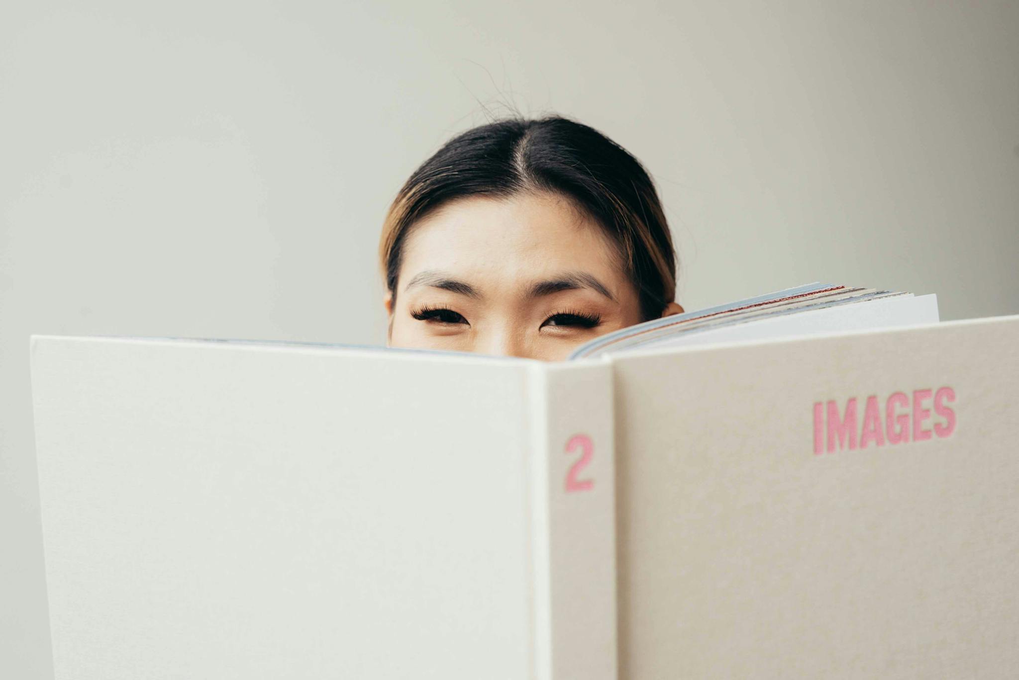 Pretty Asian woman holding opened white book with pink title while looking at camera in studio on white background